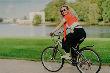 Fototapeta na wymiar Image of female model rides bicycle, looks aside with cheerful expression, wears sunglasses, breathes fresh air, poses near lake and green trees, covers long distance, finds out something new