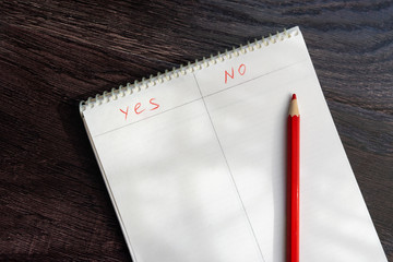A notebook with the words yes and no and a red pencil lie on a wooden surface. The concept of analysis and choice.