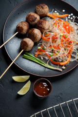 Nem nuong or fried vietnamese pork meatballs on skewers with pho noodles, elevated view on a black stone background, studio shot