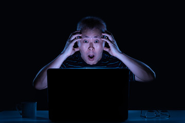 Middle age Asian male working on his computer at night in his home office, surprise to see content on his computer screen