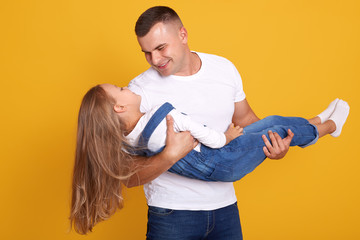 Front view of father holding little daugther wearing shirt and overalls in hands isolated over yellow studio background, handsome man dressed casual outfit looking at his charming little girl.