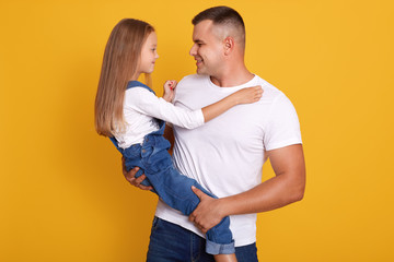 Horizontal shot of handsome father carrying his little daughter, models posing isolated over yellow background, daddy and his child looking at each other, dressed casual clothing. Family concept.