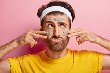 Serious male model applies cosmetic mud mask on face, wears white headband, yellow t shirt, cares...