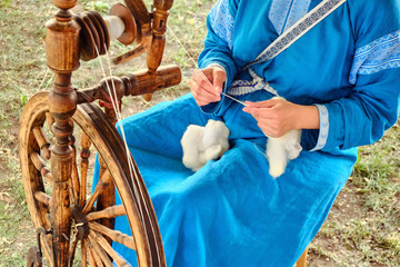 A woman in a blue dress spinning a spindle. Retro wooden wheel for thread making.