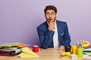 Terrified male director stares with bugged eyes, wears spectacles and elegant suit, holds chin,...