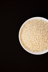 Organic Arborio rice in a white bowl on a black surface, top view. Flat lay, overhead, from above. Copy space.