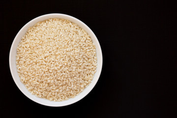 Organic Arborio rice in a white bowl on a black background, top view. Flat lay, overhead, from above. Space for text.