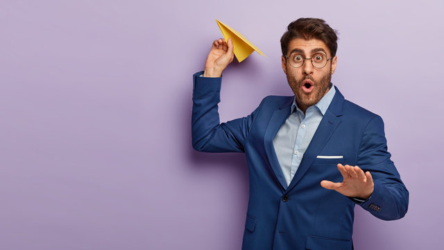 Image of prosperous busiessman holds paper aircraft, going to launch it, wears round spectacles and formal blue suit, poses against purple background with blank space. Shocked male entrepreneur