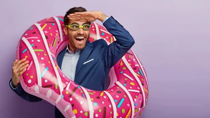 Foto op Plexiglas Photo of overjoyed male office worker focused into distance, keeps palm near forehead, pretends being on beach, wears goggles for swimming, poses in inflated swimring, notices something funny © Wayhome Studio