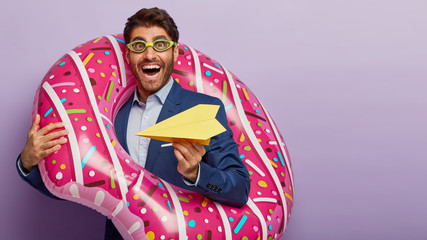 Fototapeta premium Horizontal view of happy businessman throws handmade paper plane, being successful director, carries donut ring, wears goggles for diving, formal outfit, stands agaist purple wall, free space aside