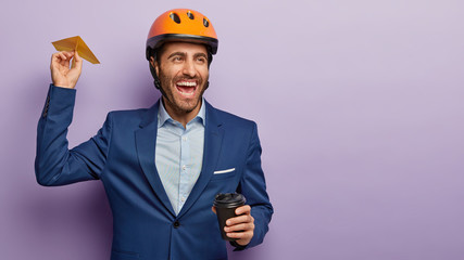 Image of happy carefree male engineer throws handmade plane, drinks coffee, glad to achieve success at work, looks into distance and laughs, models against purple wall with free space for advert