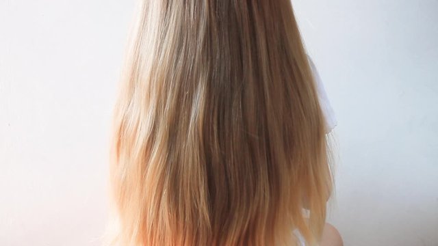 Cute girl with long blond hair, back view on light background. Female hand holds a comb and makes hairstyle. Mother and daughter.A child in a white T-shirt is sitting on a chair in hairdressing salon.