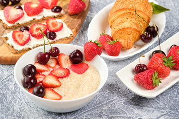 white bowl of porridge with strawberries and cherries, croissants and crispbread with cheese and strawberries on a gray background