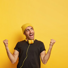 Overjoyed man raises clenched fists, feels energized and upbeat, wears yellow hat and black t...