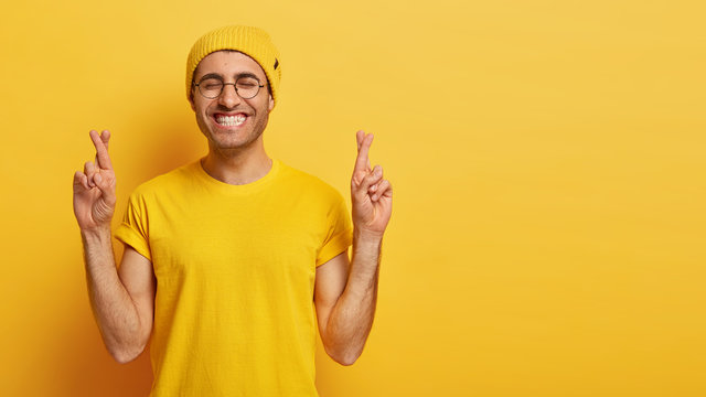 Horizontal shot of happy young man believes in good luck and fortune, keeps fingers crossed, smiles joyfully, keeps eyes shut, wears yellow outfit, poses in studio. People, body language, wish concept