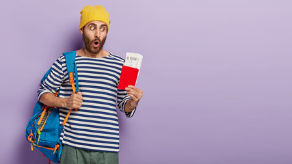 Shocked hipster guy holds passport and flying tickets, carries rucksack, ready for flight and journey abroad, dressed in casual wear isolated over purple background with blank space. Traveling concept