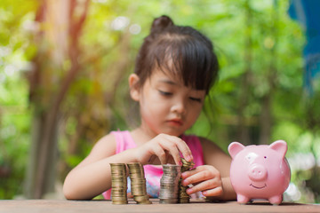 cute little girl holding coin of money and put in pink piggy bank with blur background. subject is...