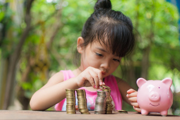 cute little girl holding coin of money and put in pink piggy bank with blur background. subject is...