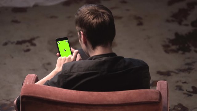 Top view of young man with brown hair sitting in chair and tapping on iPhone green screen in dark room. Stock footage. Chroma key