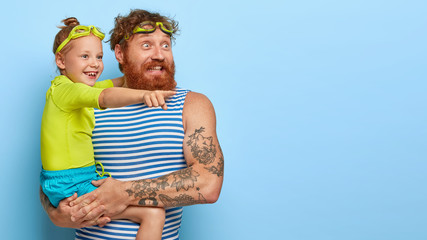 Happy surprised redhaired bearded man stands with daughter, look glafully into distance, wear...