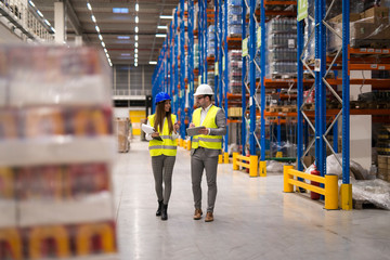 Warehouse workers walking through large storage department discussing about distribution and...