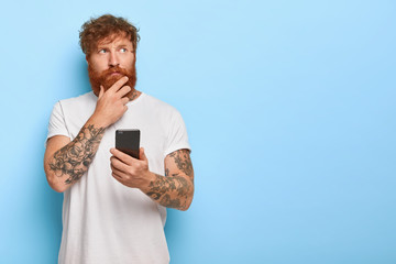 Horizontal shot of serious contemplative adult man touches thick red beard, holds mobile phone,...