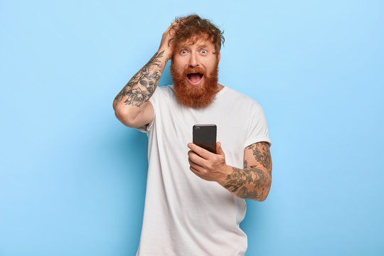 Frustrated stressful ginger hipster keeps hand on head, looks with worried facial expression, opens mouth, holds modern cellular, feels fear because of making mistake, did something wrong with app
