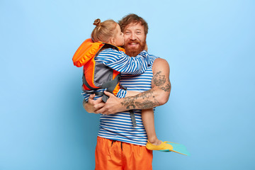 Glad father happy to get affectionate kiss and hug from daughter, holds her on hands, dressed in...