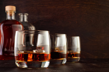 Three glasses of whiskey and ice on the wooden table. In the background is a full bottle of whiskey and open.