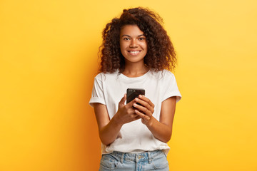 Happy smiling woman holds mobile phone, texts message on cellular, surfes internet, has curly bushy hairstyle, dressed in white casual t shirt and jeans, poses against yellow wall. Lifestyle concept