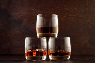 Three glasses of whiskey and ice stand in a pyramid on a wooden table.