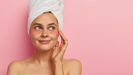 Studio shot of good looking satisfied woman with beauty pads under eyes, has daily spa procedures, healthy fresh skin, looks with green eyes aside, stands against pink background, free space aside