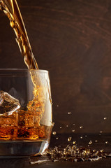 A glass of whiskey with ice on the wooden table. Top in a glass filled with whiskey. Right splashes from the drink.