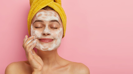 Close up portrait of satisfied beautiful woman massages face, washes with soap, poses bare shouldes, has healthy smooth skin, keeps eyes closed, isolated over pink background free space for your promo