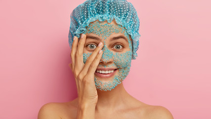 Image of lovely satisfied beautiful woman looks through fingers, has blue sea salt granules on face, does peeling procedure for skin, smiles pleasantly, wears protective headgear after taking shower