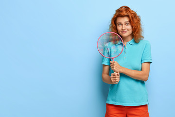 Pretty ginger woman with curly hair, likes tennis, holds racket, ready for playing, wears casual summer outfit, poses against blue wall, copy space on left side for your advert. People and lifestyle
