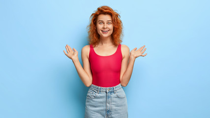 Positive good looking girl with ginger hairstyle, spreads hands, gets awesome surprise, has toothy smile, dressed in red top and denim shorts, isolated over bluue studio wall. Emotions concept