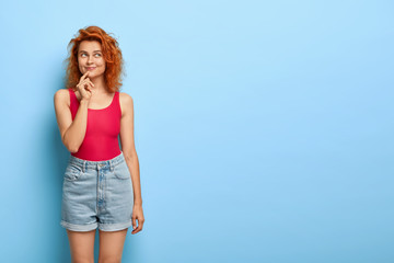 Indoor shot of happy woman with ginger hair, has thoughtful expression, dressed in casual vest and denim shorts, imagines something pleasant, isolated over blue studio wall, copy space area.