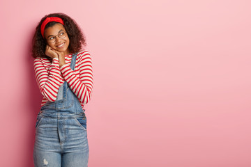 Studio shot of happy millennial Afro American girl looks happily aside, wears red headband, striped jumper and denim overalls, poses against pink background with copy space for your promotion