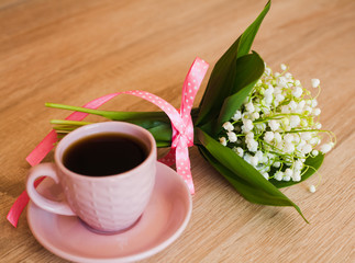 Bouquet of beautiful lilies of the valley tied with a pink ribbon and cup of coffee on wooden table. Close-up.