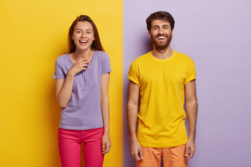 Photo of two delighted young woman and man stand together, express good emotions, smile happily, spend free time together, have fun, pose against yellow and purple background, dressed casually