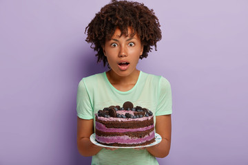 Photo of surprised dark skinned woman with curly haircut, holds delicious cake, surprised guests...