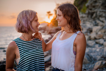 Two young cheerful women in hipsters hats on a rock on the coast of the sea. Summer landscape with girl, sea, Islands and orange sunlight.