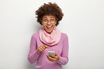 Pleasant looking female with crisp hair, points at cell phone device, donwloads new modern...