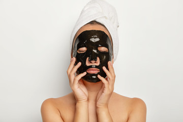 Close up shot of serious young woman wears black facial mask, has eyes closed, touches face gently, wears wrapped towel around head, poses against white background. Shirtless female model indoor