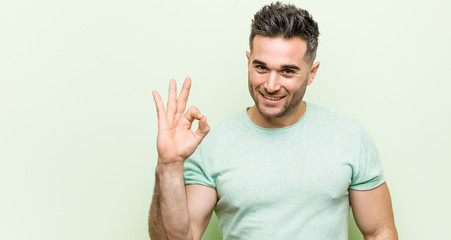 Young handsome man against a green background cheerful and confident showing ok gesture.