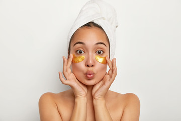 Photo of lovely female model applies yellow pads under eyes for reducing wrinkles, has anti aging procedures, keeps lips folded, stands shirtless indoor, wrapped towel on head. Cosmetology concept