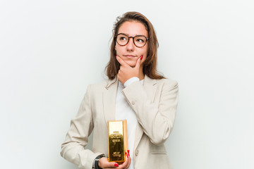 Young european business woman holding a gold ingot looking sideways with doubtful and skeptical...