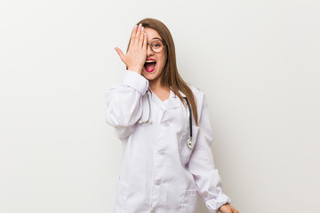 Young doctor woman against a white wall having fun covering half of face with palm.