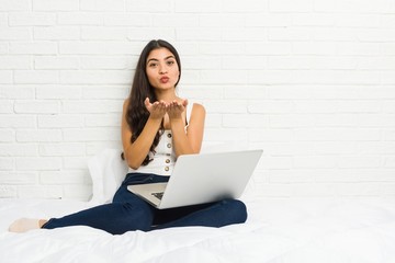 Young arab woman working with her laptop on the bed folding lips and holding palms to send air kiss.